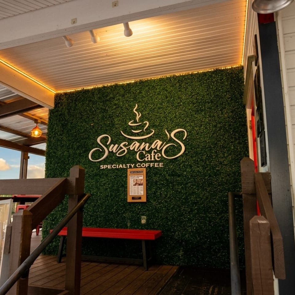 Susana’s Cafe entrance featuring a greenery wall, specialty coffee sign, wooden deck, red seating, and menu board.