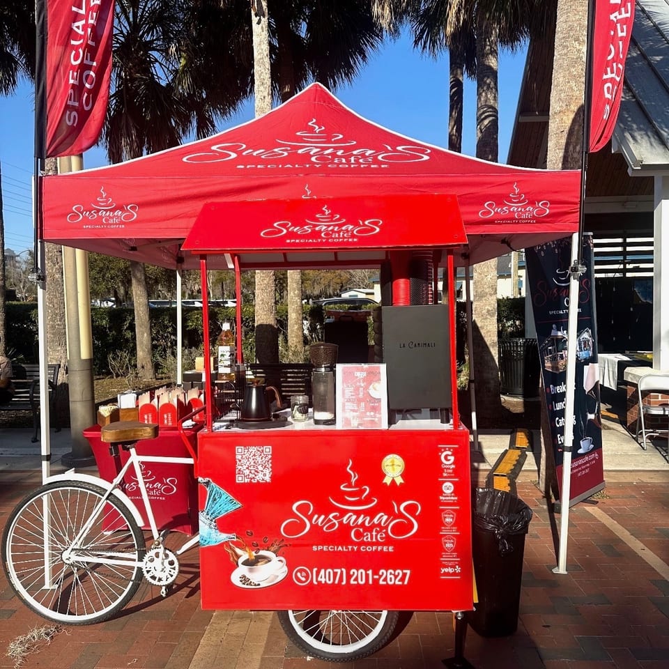 Susana's Cafe red coffee cart with a matching tent and flags under palm trees.