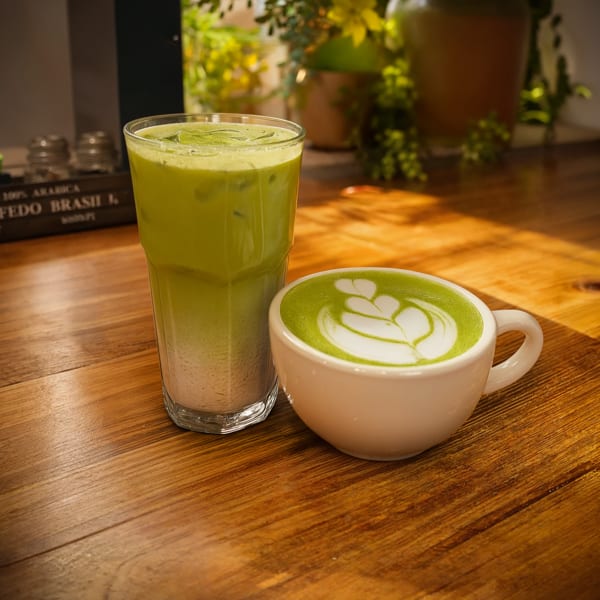 Green iced drink and matcha latte with heart-shaped foam art on a warmly lit wooden café table.