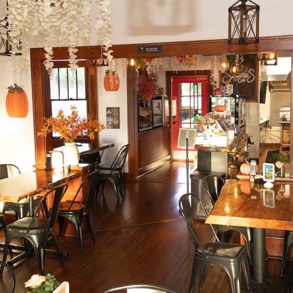 Warm café interior with wooden tables, black metal chairs, floral decorations, pumpkins, and a coffee counter.
