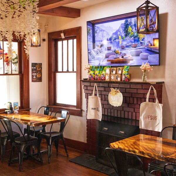 Warm restaurant dining room with polished wooden tables, black chairs, brick fireplace, decorative flowers, and mountain-lake artwork.