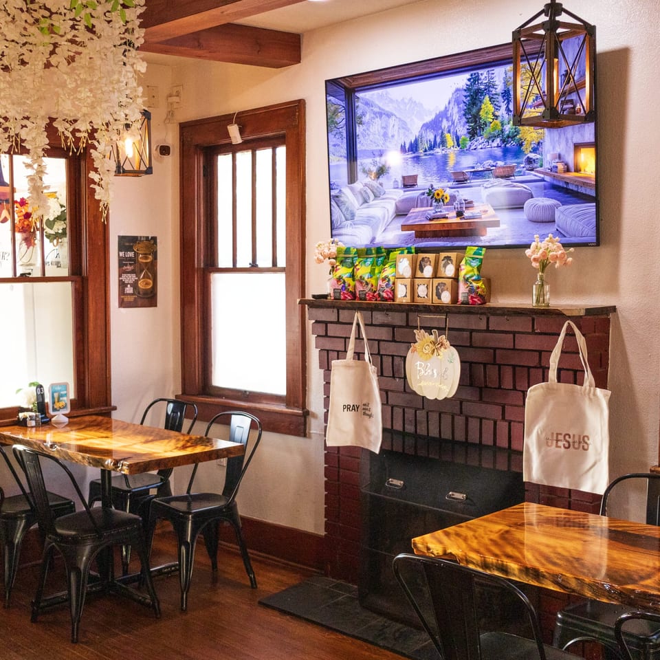 Warm restaurant dining room with wooden tables, black chairs, brick fireplace, hanging flowers, and mountain scenery displayed on television.