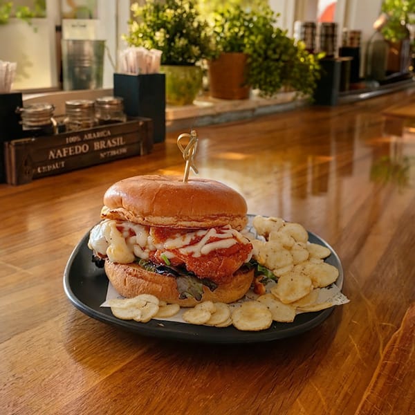 Layered gourmet burger with sauce and greens, surrounded by potato chips on a black plate atop a wooden restaurant table.