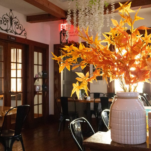 Warmly lit restaurant interior with wooden tables, black chairs, decorative lanterns, and a vase of golden autumn leaves.