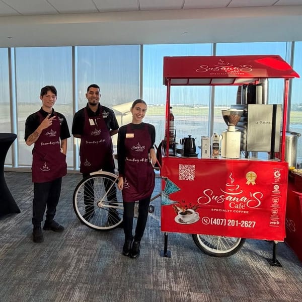 Three café staff pose beside Susana’s Café red specialty coffee cart in a bright airport-like space.