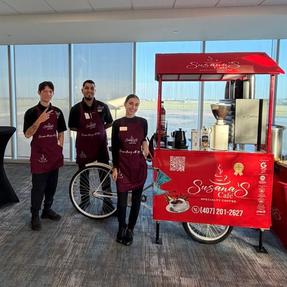 Three café staff pose beside Susana’s Café red specialty coffee cart in a bright airport-like space.