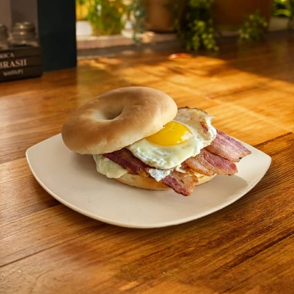Bagel sandwich with bacon, melted cheese, and a fried egg served on a white plate atop a wooden table.